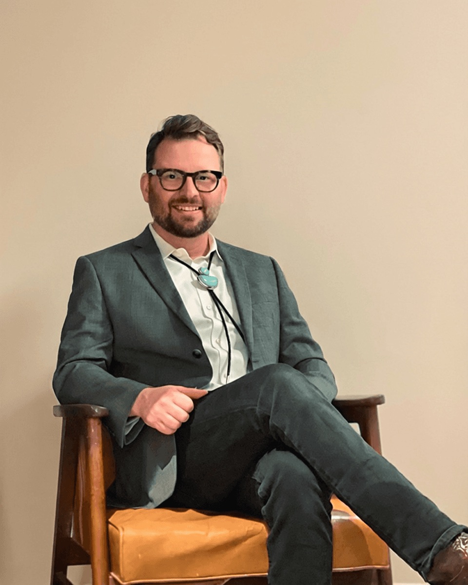 George Irwin, seated in a wooden chair, in a gray sport coat and turquoise bolo tie.